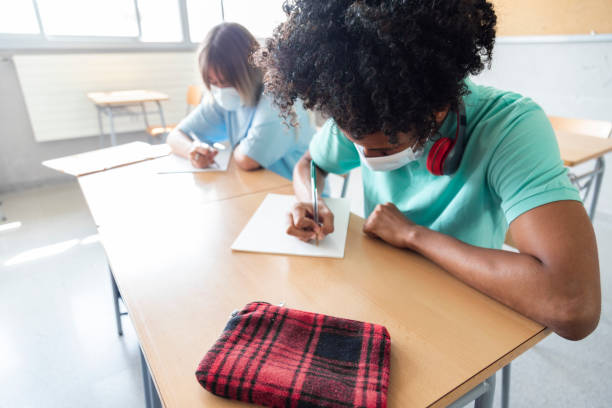 Black teen boy wearing face mask in class. High school student writing on paper. Copy space. Educaction concept. Healthcare concept.