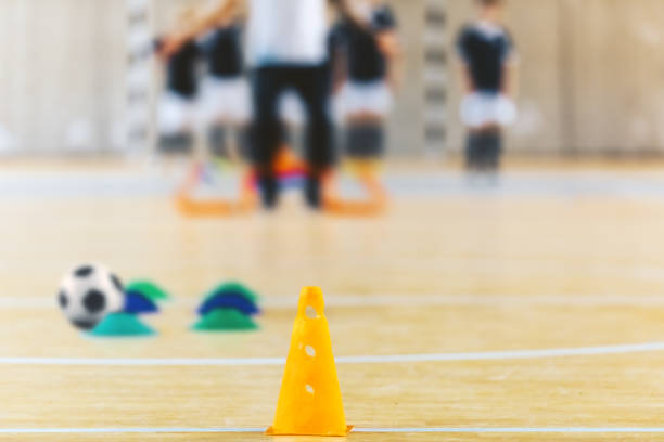 Group of kids training soccer at indoor soccer field. Indoor football training arena. Coach training with kids on wooden floor. Indoor soccer practice equipment; yellow cone and training markers
