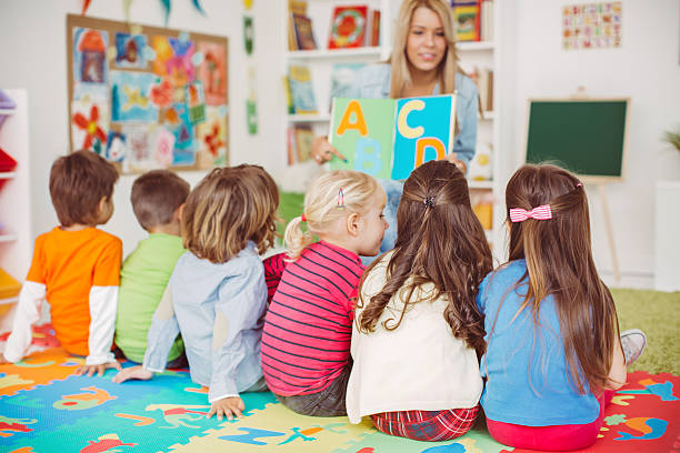 Teacher with a group of preschool children in a nursery. The children are sitting on the floor and listening  teacher. Learning letters. In the background we can see a shelf with some, toys, black board and books. View from behind.